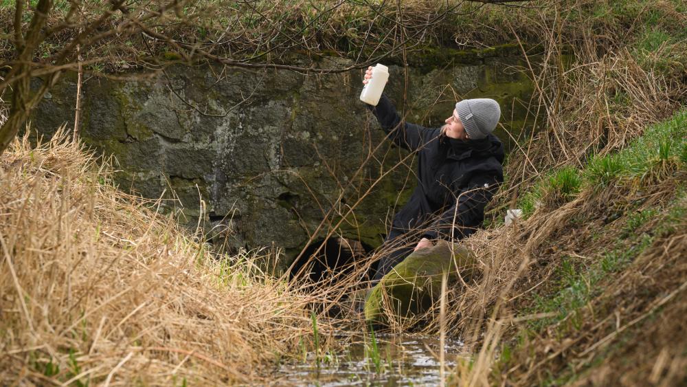 Eine Technikerin nimmt eine Wasserprobe an einem Graben..