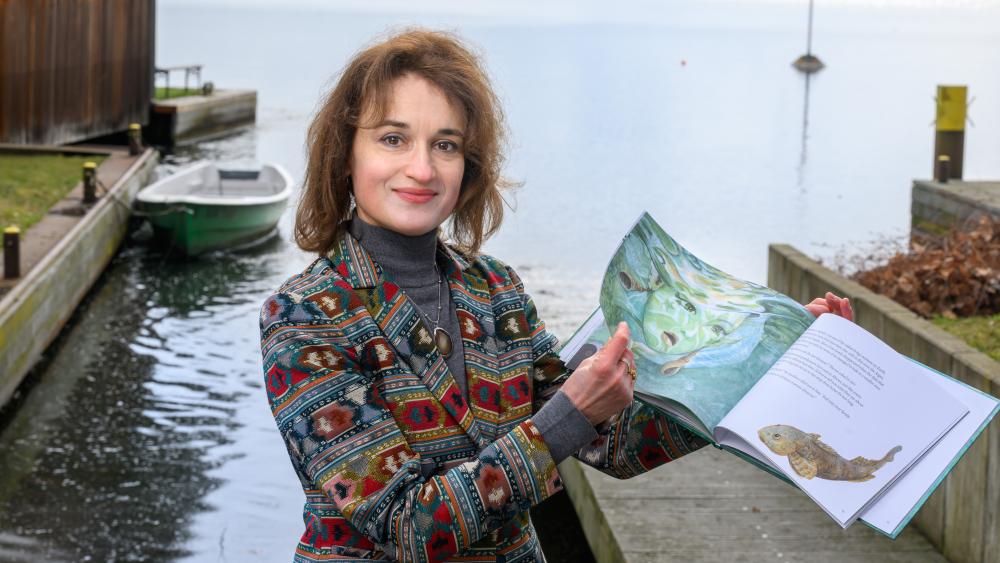 Gabriela Costea steht vor dem Müggelsee und hält das Kinderbuch geöffnet in der Hand.