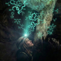 Eine Person mit Schutzhelm und Stirnlampe steht im Profil in einer dunklen H&ouml;hle und blickt nach oben zu einer Felsformation. Digital &uuml;berlagerte, leuchtend t&uuml;rkisfarbene Grafiken von DNA-Doppelhelices und chemischen Molek&uuml;lstrukturen schweben &uuml;ber der Person und scheinen aus dem Gestein zu entspringen, was wissenschaftliche Entdeckung symbolisiert.